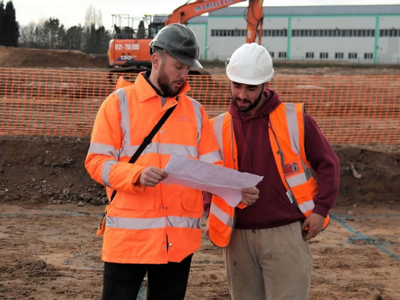 Two men wearing hard hats in hi-vis clothing looking at a plan